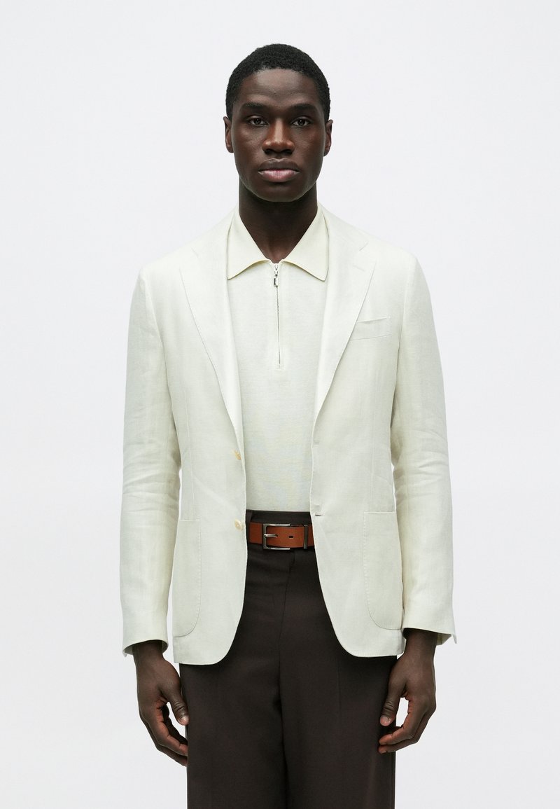 Homme portant un blazer blanc cassé et une chemise à col zippée, un pantalon marron foncé avec une ceinture marron, debout devant un fond blanc uni.