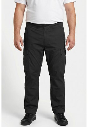 Hombre que lleva pantalones cargo negros con bolsillos laterales, una camiseta blanca y zapatillas negras, de pie frente a un fondo sencillo.