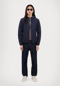 Ναυτικά κ κουβερτίνα μπουφάν με κουκούλα, φερμουάρ, γκρι μπλούζα, σκούρα τζιν παντελόνια και λευκά αθλητικά παπούτσια. Μοντέλο που φοράει γυαλιά ηλίου.