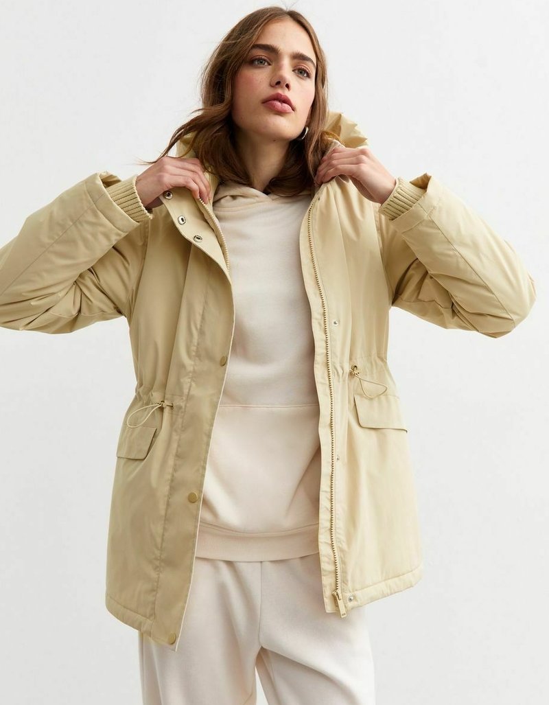 Light beige padded jacket with a hood, elastic cuffs, drawstring waist, and snap fasteners. Worn over a cream hoodie.