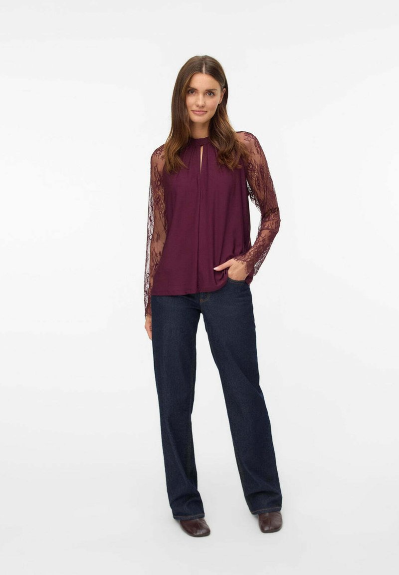 Blusa bordeaux con collo a chiave, corta davanti e maniche lunghe in pizzo trasparente, abbinata a jeans svasati blu scuro e scarpe marroni.