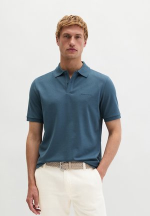 Homme portant un polo à manches courtes bleu sarcelle avec une ceinture tissée beige et un pantalon blanc, posé devant un fond blanc uni.