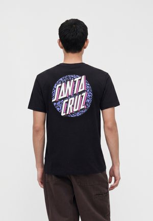 Hombre con una camiseta negra con un gran logo "Santa Cruz" en púrpura y rosa en la espalda, de pie frente a un fondo claro y liso.