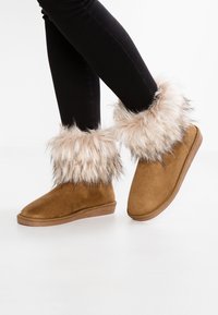 Botines de ante marrón con un suave puño de pelaje sintético de color beige claro, que presentan una suela plana y un detalle de costura texturada alrededor de la base.