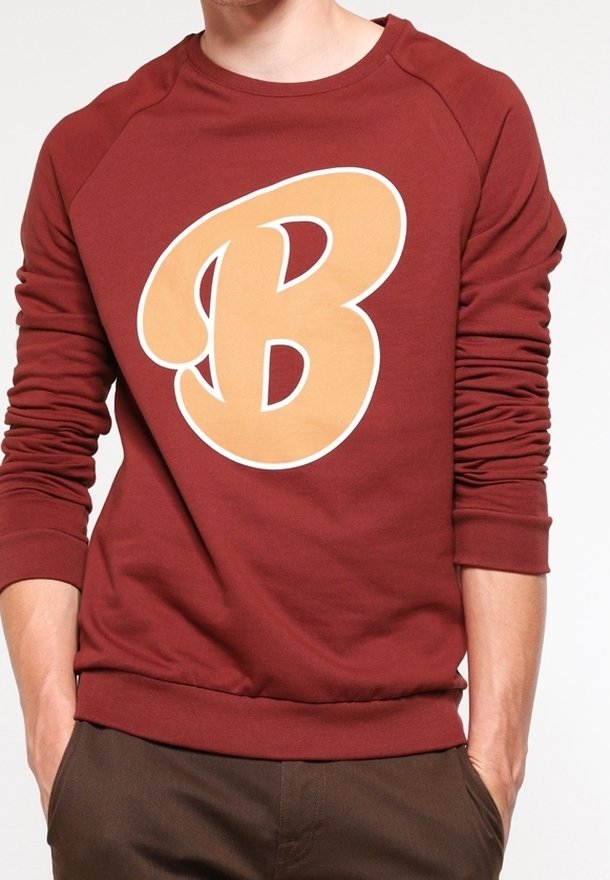 Personne portant un sweat-shirt bordeaux à manches longues avec une grande lettre beige « B » sur le devant, mains dans les poches, cadré au niveau du cou.
