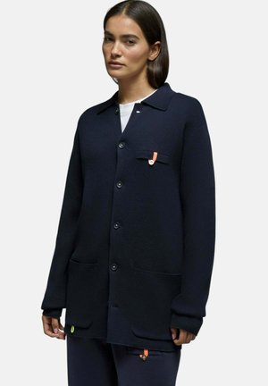 Femme portant un cardigan bleu marine boutonné avec poches avant et détails orange, associé à un pantalon bleu marine assorti sur un fond uni.