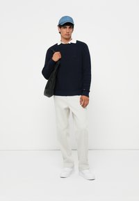 Marine gebreid sweater, witte kraagbloes eronder, lichtgrijze broek, zwarte tas, blauwe pet en witte sneakers op een effen achtergrond.