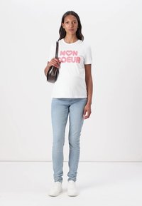 Balta trumpa rankovė t-shirt su rožiniu "MON COEUR" užrašu, derinama su šviesiai mėlynomis džinsomis ir baltais sportbačiais. Modelis laiko rudą maišą.