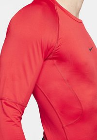 Rød atletisk langærmet top lavet af strækstof med buede sømme og et lille sort Nike-logo på brystet. Glat tekstur.