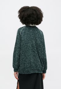 Pólo oversized de polar verde escuro com um acabamento texturizado, apresentando bainhas brancas ao longo dos ombros e punhos elásticos.