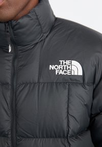 Kadar u bližem planu prikazuje osobu u crnoj jakni s preklopom i visokim ovratnikom, s logotipom The North Face na prsima.