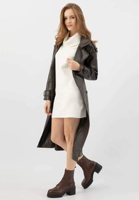 Cappotto lungo in pelle marrone con vita accentuata da una cintura, indossato sopra un vestito bianco, abbinato a stivaletti alla caviglia marroni con lacci. Tessuto liscio, vestibilità su misura.