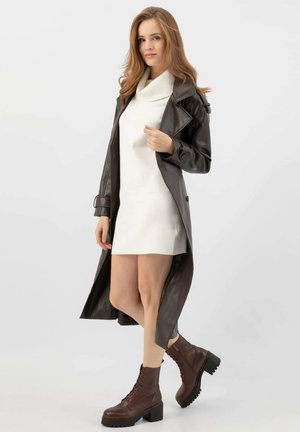 Manteau long en cuir marron avec une taille ceinturée, superposé sur une robe blanche, assorti à des bottines à lacets marron. Texture lisse, coupe ajustée.