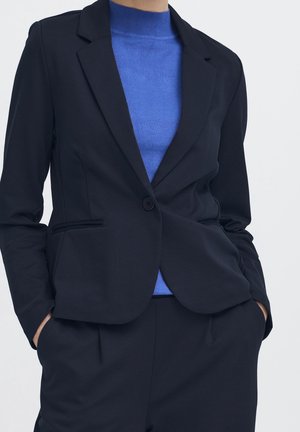 Vrouw draagt een op maat gemaakte zwarte blazer en bijpassende broek over een blauwe coltrui, met handen in de zakken tegen een effen achtergrond.