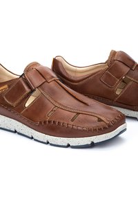 Zapatos de cuero marrón sin cordones, con detalles recortados, un diseño cosido y una suela blanca texturizada con acentos negros.