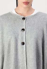 Cardigan cinza de lã com decote redondo, costura branca decorativa e três grandes botões pretos na frente.