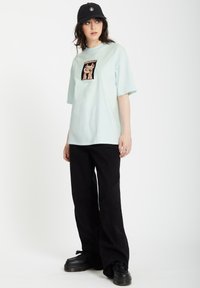 Hellblauses T-Shirt mit kurzen Ärmeln und grafikbedrucktem Patch auf der Vorderseite, kombiniert mit schwarzen weiten Hosen und schwarzen Plateau-Schuhen. Inklusive lässiger Kappe.