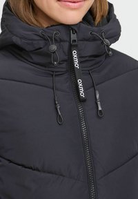 Veste rembourrée noire avec un col haut, fermeture éclair à l'avant et cordons de serrage réglables. Présente une texture matelassée et un tirant de fermeture éclair estampillé.