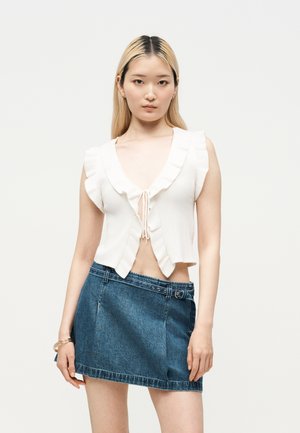 Giovane donna con lunghi capelli biondi che indossa un top bianco senza maniche con volant e una minigonna di jeans blu con cintura, in piedi davanti a uno sfondo uniforme.