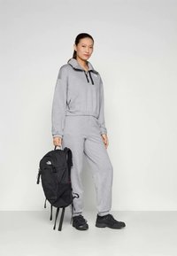 Pullover cinza de fleece com um fecho de correr preto e logótipo, combinado com calças de agasalho cinzas. Uma mochila preta é segurada numa mão. Sapatos de caminhada pretos completam o visual.