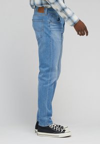Jeans en denim bleu clair avec un design ajusté, une pièce en cuir beige à l'arrière et des poches arrière. Associés à des baskets montantes noires.