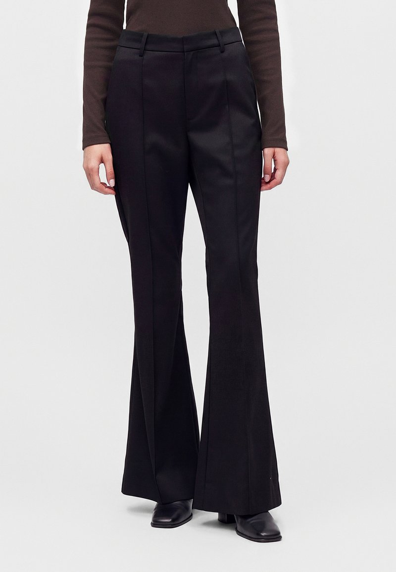 Personne portant un pantalon noir taille haute à jambes larges avec un pli frontal, associé à des chaussures noires et un haut marron à manches longues.