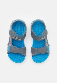 Blå og grå sandaler med teksturerte gummisåler, justerbare stropper og et diamantmønster på innersålen. Merket viser "SKECHERS SHARK-BOTS."