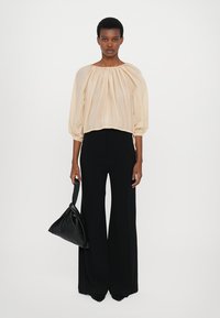 Leichte beigem Bluse mit Puffärmeln und gerafftem Ausschnitt, kombiniert mit hoch-taillierten, schwarzen, weiten Hosen. Das Model hält eine schwarze Tasche.