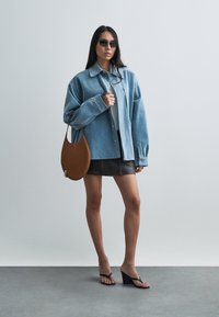Woman wearing oversized light blue denim jacket, dark mini skirt, black wedge sandals, sunglasses, and carrying a brown leather shoulder bag.