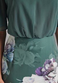 Robe fleurie avec un haut fluide et transparent de couleur verte, cintrée à la taille, et une jupe ajustée ornée de grands motifs floraux violets et blancs.