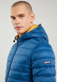 Mann mit kurzen Haaren, der eine blaue gesteppte Jacke mit gelber Kapuzenfütterung und dem "Big Star"-Logo auf dem linken Ärmel trägt.
