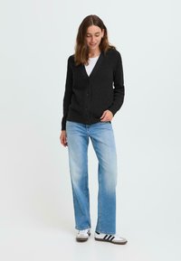 Cardigan noir côtelé avec un col en V et une fermeture à boutons, associé à un jean évasé bleu clair et des baskets blanches avec des rayures noires.