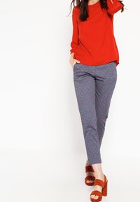 Červený top s dlouhými rukávy v kombinaci s modrými kalhotami se vzorem s geometrickým designem. Modelka má na sobě hnědé platformové sandály.