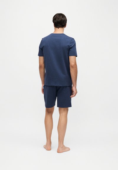 Homme debout pieds nus, dos à la caméra, portant une chemise à manches courtes bleu foncé et un short assorti sur un fond clair uni.