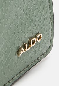 Grön texturerad läderyta med guldmetalliska ALDO-bokstäver och synlig sömnad längs kanten.