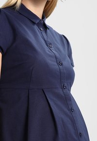 Tmavě modré šaty na knoflíky s límečkem, krátkými rukávy a plisovaným designem. Vyrobené z texturované látky s viditelnými knoflíky.
