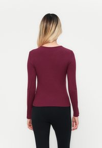 Tops en rib avec manches longues de couleur bourgogne, à col rond et à poignets évasés, fabriqué à partir d'un tissu doux et texturé. Associé à un pantalon noir ajusté.