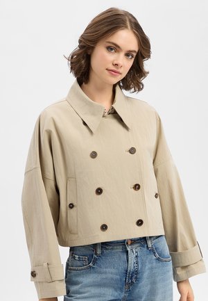 Ung kvinde med kort krøllet hår iført en beige cropped dobbeltradet jakke og blå jeans, stående fremad med et neutralt udtryk.