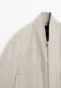 Beige Wolljacke mit hohem Kragen, Frontreißverschluss und zwei Seitentaschen. Weiche Textur mit dezentem, neutralem Farbton.