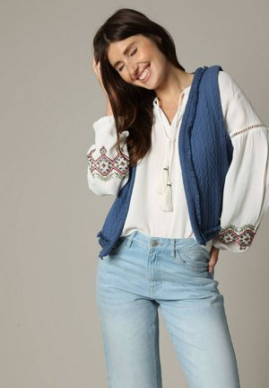 Gilet matelassé bleu sur une blouse blanche avec des manches longues brodées, associé à un jean bleu clair. Comprend des détails à franges sur la blouse.
