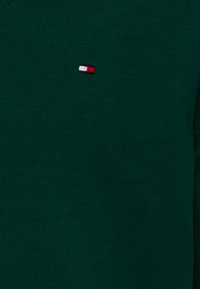 Tissu tricoté vert foncé avec un petit logo rectangulaire aux couleurs blanc, bleu marine et rouge.