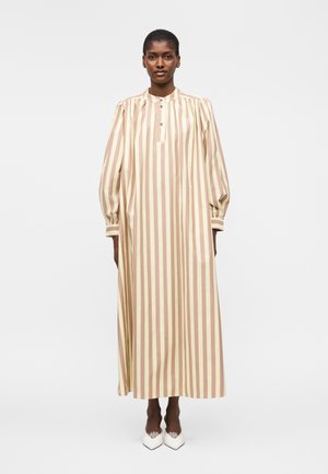 Vrouw draagt een lange beige en lichtbruine jurk met verticale strepen, lange opgeblazen mouwen en witte puntige schoenen, staand tegen een witte achtergrond.