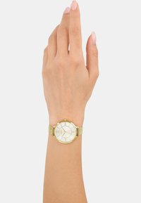 Goldene Armbanduhr mit einem weißen runden Ziffernblatt, strukturiertem goldenen Mesh-Armband und römischen Ziffern als Stundenmarkierungen. Elegantes und schlichtes Design.