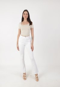 Jeune femme aux longs cheveux bruns, portant un top beige à encolure carrée, un jean flare blanc taille haute et des sandales à talons beiges, debout.