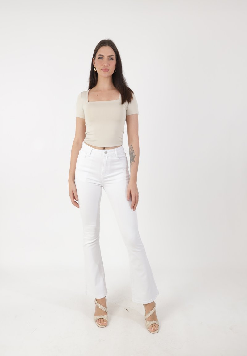 Jeune femme aux longs cheveux bruns, portant un top beige à encolure carrée, un jean flare blanc taille haute et des sandales à talons beiges, debout.