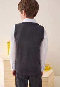 Veste grise en tricot sans manches, avec encolure et ourlet côtelés, portée par-dessus une chemise blanche à manches longues, vue de dos.