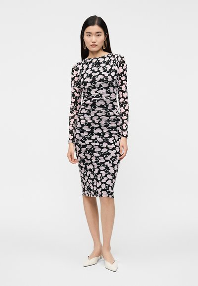 Vestido comprido preto de manga comprida com padrão floral em rosa e branco, apresentando detalhes franzidos e comprimento midi ajustado. Sapatos brancos de bico fino.