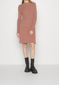 Robe en maille côtelée couleur rose poudré avec manches longues, col montant et boutons dorés sur les épaules. Associée à des bottes noires épaisses.