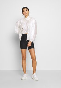 Veste bomber irisée au design épuré, associée à un top blanc noué, un short noir et des baskets blanches. Look simple et moderne.
