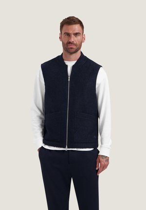Man met kort haar die een wit shirt met lange mouwen draagt onder een marineblauw vest met rits en donkere broek, staand met handen in de zakken.